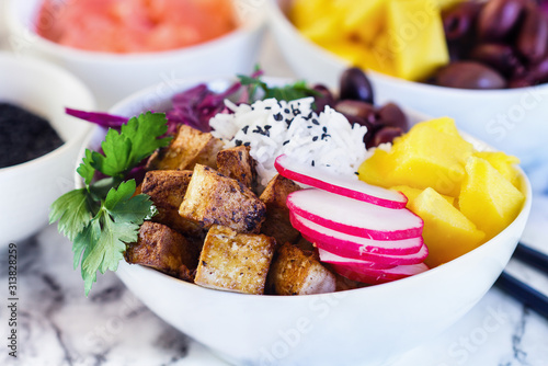 Healthy vegan poke bowl or Buddha bowl with basmati rice, mango, fried tofu, purple cabbage, radishes, kalamata olives, pickled ginger and black sesame seeds. Focus on tofu with blurred background.