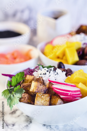 Healthy vegan poke bowl or Buddha bowl with basmati rice, mango, fried tofu, purple cabbage, radishes, kalamata olives, pickled ginger and black sesame seeds. Focus on tofu with blurred background.