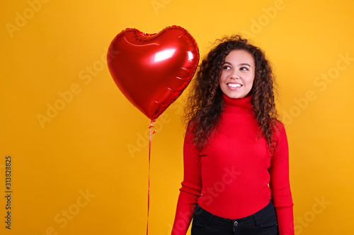 Studio portrait of young woman with dark skin and long curly hair wearing sexy dress over the festive red wall with heart shaped balloon. Close up, isolated background, copy space. © Evrymmnt
