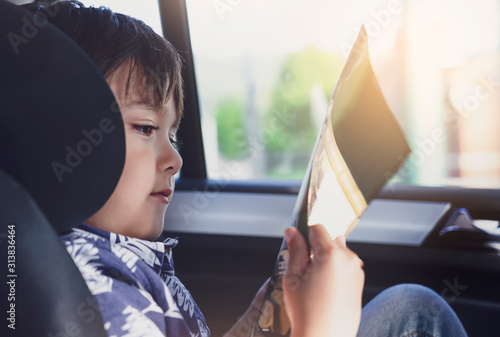 Kid siting on car seat and reading a book, Little boy sitting in the car in child safety seat, Portrait of toddler entertaining himserf on a road trip. Concept of safety taveling by car with children photo