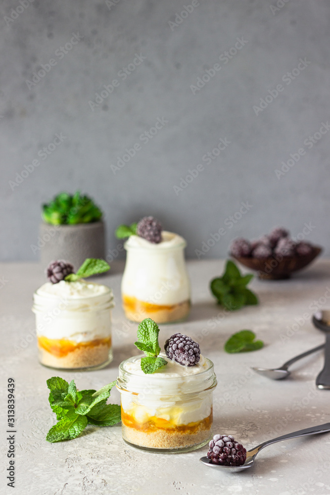 Homemade no baked dessert (cheesecake, trifle, mousse) with orange or peach jam in small portioned jars served with frozen berries and mint. Light grey stone background, copy space. Homemade no baked dessert (cheesecake, trifle, mousse) with orange or peach jam in small portioned jars served with frozen berries and mint. Light grey stone background, copy space.