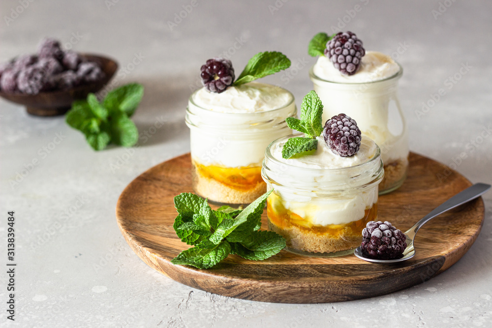 Homemade no baked dessert (cheesecake, trifle, mousse) with orange or peach jam in small portioned jars served with frozen berries and mint. Light grey stone background, copy space. Homemade no baked dessert (cheesecake, trifle, mousse) with orange or peach jam in small portioned jars served with frozen berries and mint. Light grey stone background, copy space.