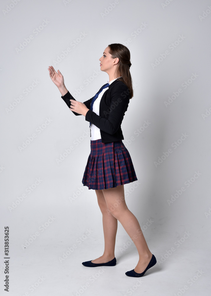 full length portrait of a pretty brunette girl wearing a school uniform of black jacket and plaid skirt. Standing pose  against a studio background.