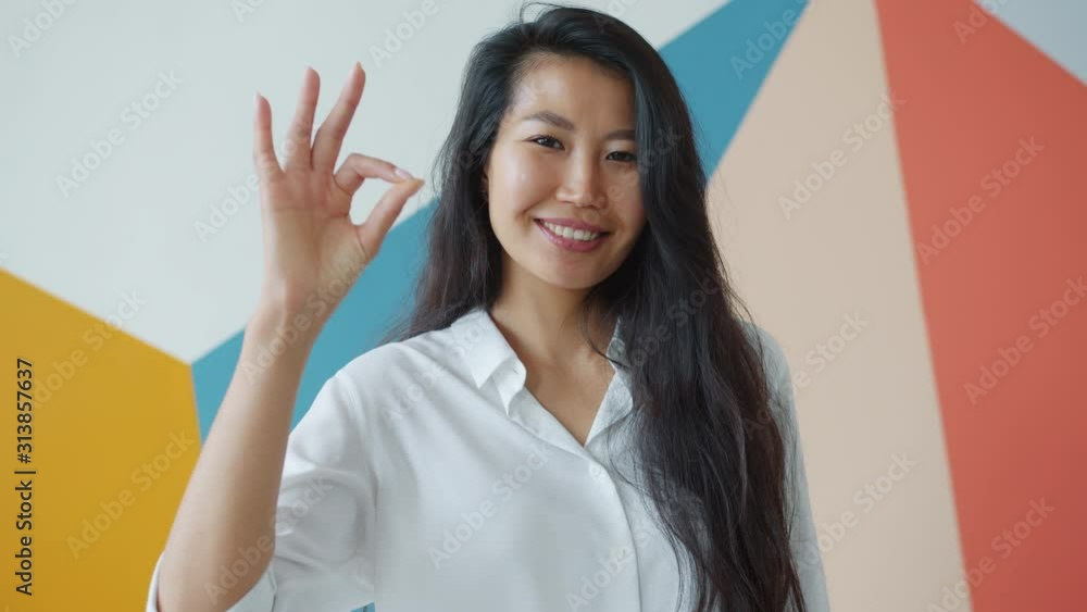 Portrait of cute Asian woman student showing okay hand gesture expressing happiness standing indoors on colorful background. People and lifestyle concept. Portrait of cute Asian woman student showing okay hand gesture expressing happiness standing indoors on colorful background. People and lifestyle concept.