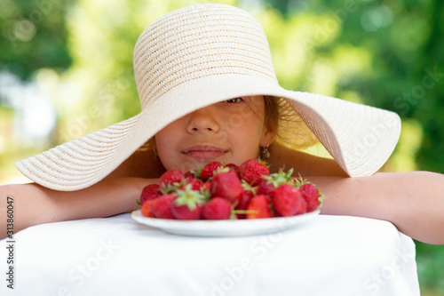 Portrait of Happy cute preteen girl in big hat is eating strawberries at summer day