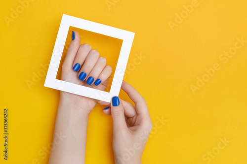 Female hands with manicure in trendy classic blue color holding white frame on the yellow background