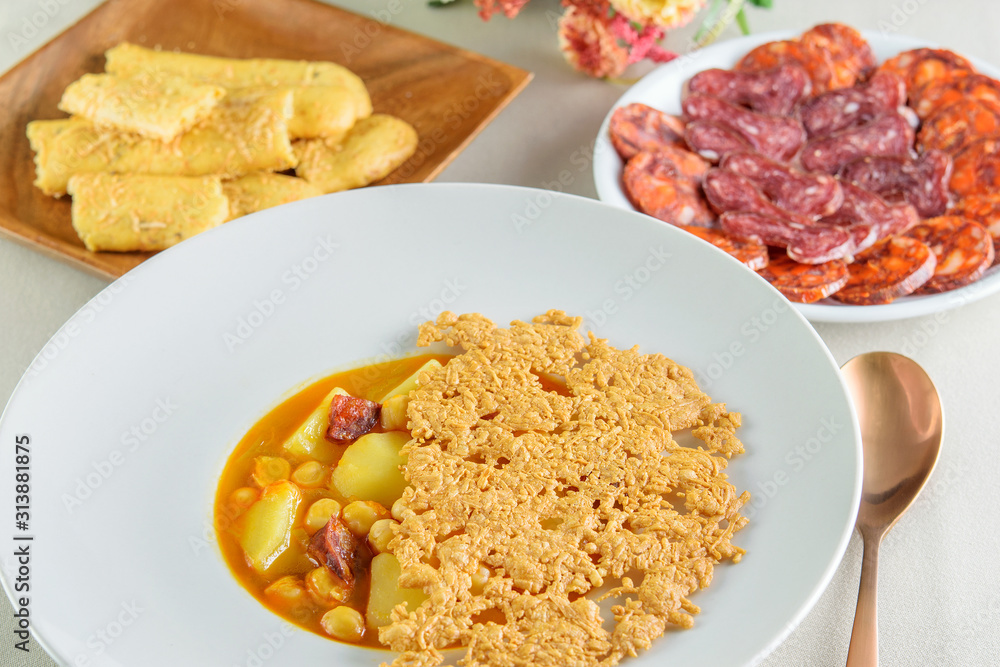 White modern bowl with fresh hot soup sauce of soybeans, potatoes and slices of sausage. Plates with slices of various sausages and cheese sticks. Healthy food prepared for eating. White modern bowl with fresh hot soup sauce of soybeans, potatoes and slices of sausage. Plates with slices of various sausages and cheese sticks. Healthy food prepared for eating.