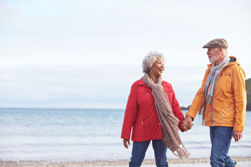 Senior Couple Hold Hands As They Walk Along Shoreline On Winter Beach Vacation