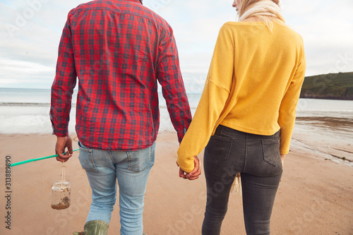 Rear View Of Loving Couple Holding Hands Walking Along Beach Shoreline On Winter Vacation