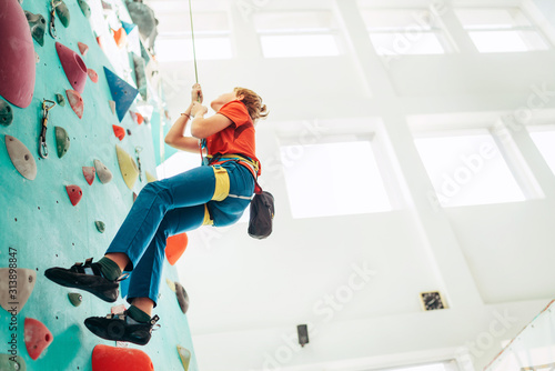 Teenager boy at indoor climbing wall hall. Boy is climbing using a top rope,chalk bag and climbing harness. Active teenager time spending concept image.