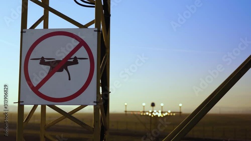 No drone zone sign on approach lighting system at runway. Airport airspace perimeter prohibition drones fly sign. Airport infrastructure and buildings in background, against blue sky.