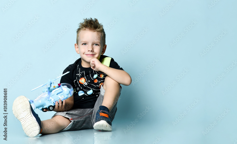 Positive blond boy in stylish casual clothing sitting on floor and holding toy helicopter present in hand over blue background Positive blond boy in stylish casual clothing sitting on floor and holding toy helicopter present in hand over blue background