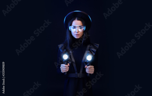 Fototapeta Naklejka Na Ścianę i Meble -  Beautiful woman in futuristic black dress over dark background. Gamer girl in glasses of virtual reality with controllers in hands. Augmented reality, game, hobby concept. VR. Blue neon light.