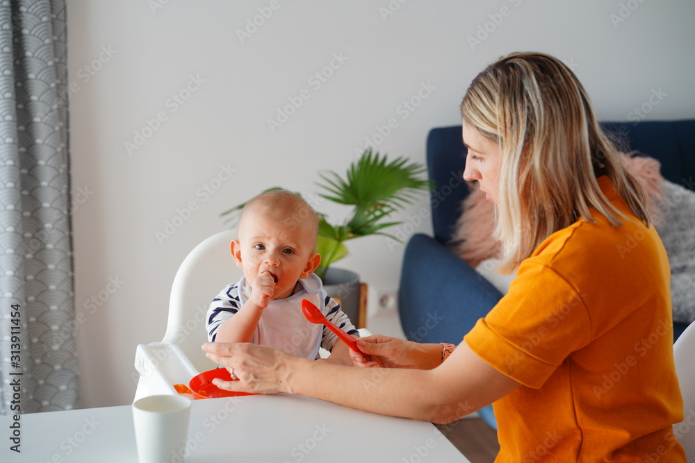 Mom feeding her baby girl with a spoon. Mother giving food to her eight-month child at home.