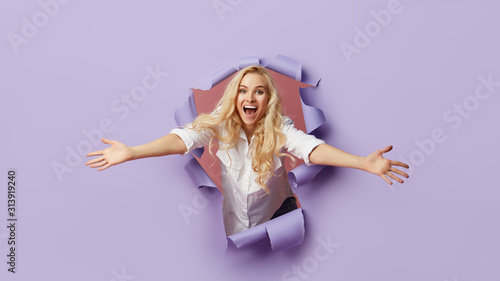Картината върху платно Surprised young women stands in torn paper hole, has excited cheerful expression, looks through breakthrough of purple background