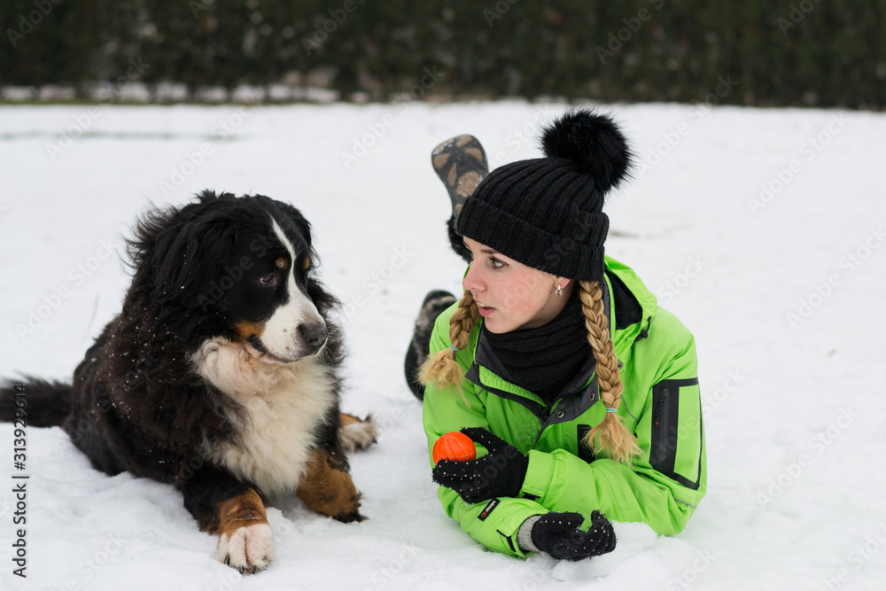 Girl is talking to her dog - berner sennenhund with the ball in hand liying on the snow in winter park. Time of friendship, happiness, and education.