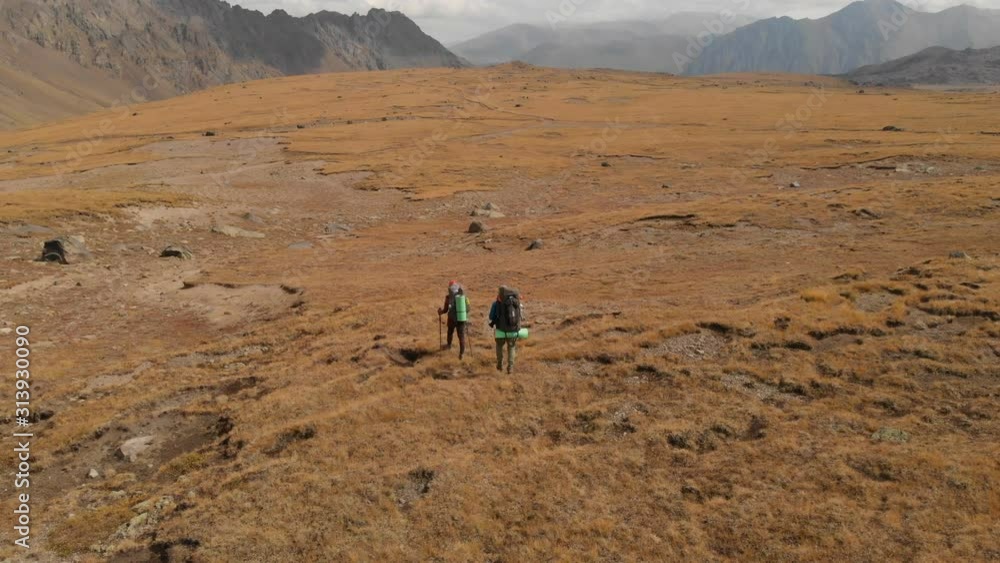 Aerial view of a couple of travelers man and woman with large backpacks in hats and sunglasses walk along an alpine plateau surrounded by epic mountains Aerial view of a couple of travelers man and woman with large backpacks in hats and sunglasses walk along an alpine plateau surrounded by epic mountains