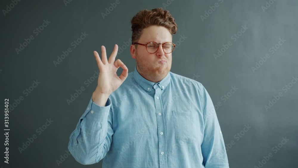 Guy with expressive face is showing ok hand gesture and winking standing alone on gray background. Body language and human positive emotions concept. Guy with expressive face is showing ok hand gesture and winking standing alone on gray background. Body language and human positive emotions concept.
