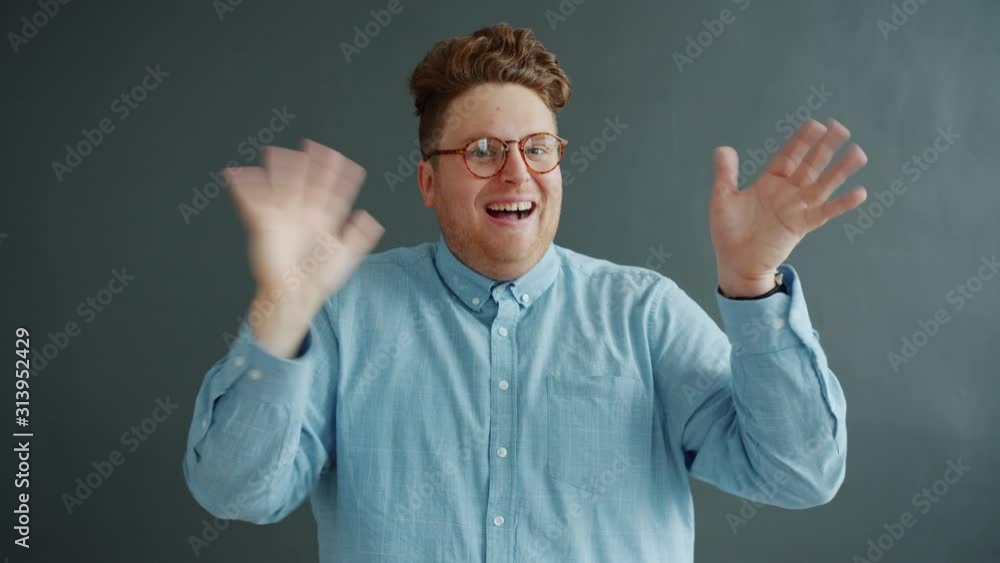 Handsome young man in glasses is waving hand with funny face standing on dark gray background alone looking at camera. People and gestures concept. Handsome young man in glasses is waving hand with funny face standing on dark gray background alone looking at camera. People and gestures concept.