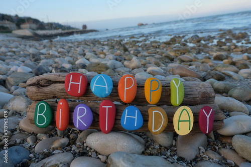 Fototapeta Naklejka Na Ścianę i Meble -  Happy Birthday text composed with multi colored stone letters on wood over a rocks beach