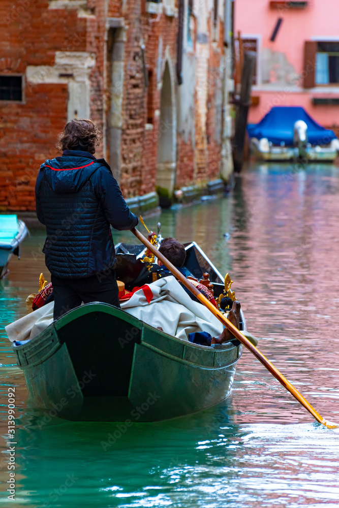 Venice, Italy. Gondolier/ Oarsman standing in gondola with single oar rowing tourists/ people. Popular romantic tourist boat ride by old brick wall gothic Venetian buildings on water canal channel.