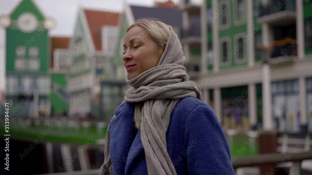the blonde is dressed in a blue coat and a gray scarf on her head walking around the old city along the green houses. she enjoys the walk, closes her eyes, smiles the blonde is dressed in a blue coat and a gray scarf on her head walking around the old city along the green houses. she enjoys the walk, closes her eyes, smiles