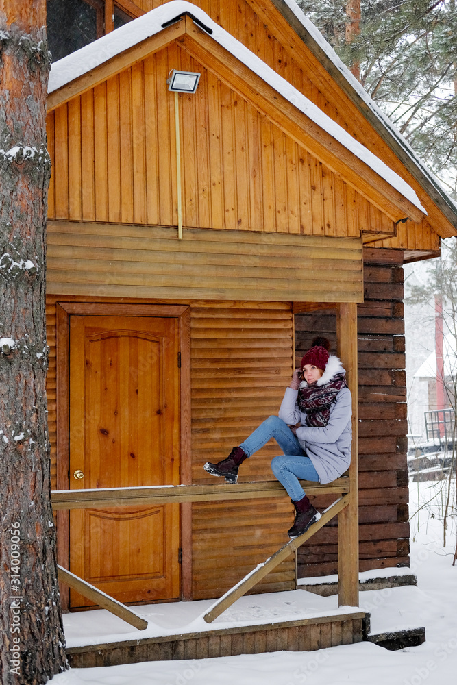 Fototapeta premium Woman sitting on snowy porch wooden rustic cottage in the winter woods. Portrait of a girl in a warm gray coat and burgundy knit scarf.