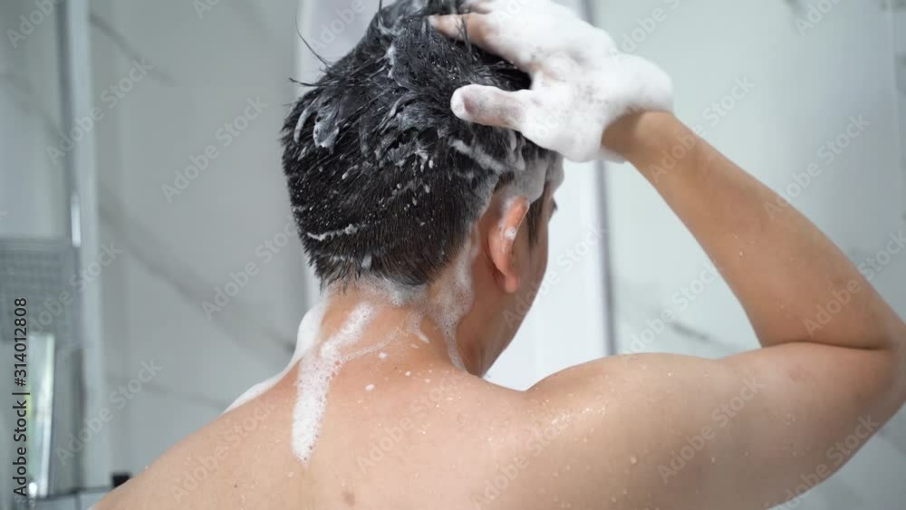 Back view from below of black haired naked man washing hair with shampoo while talking shower in bathroom Back view from below of black haired naked man washing hair with shampoo while talking shower in bathroom