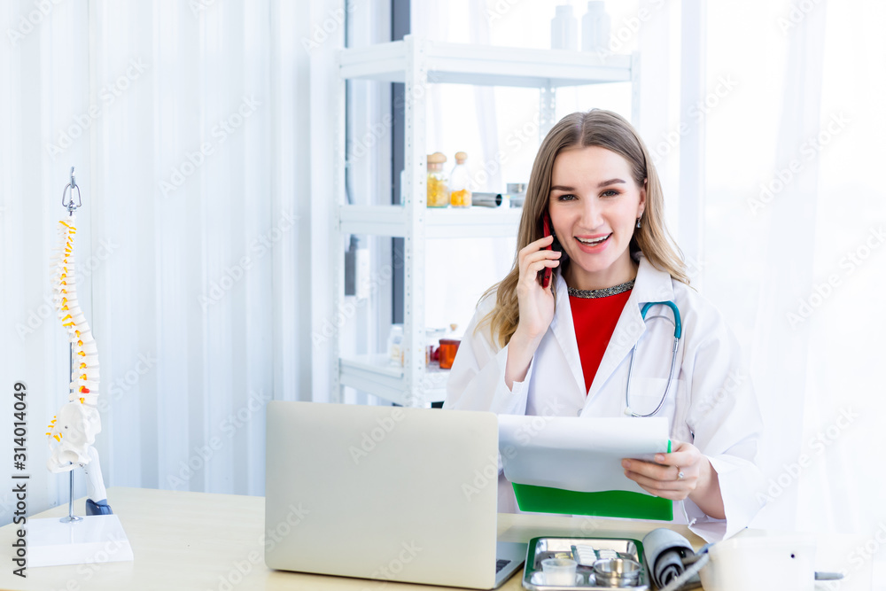 female doctor hold pick up Smartphone with stethoscope working with laptop computer and writing on paperwork on wooden table in Hospital background female doctor hold pick up Smartphone with stethoscope working with laptop computer and writing on paperwork on wooden table in Hospital background