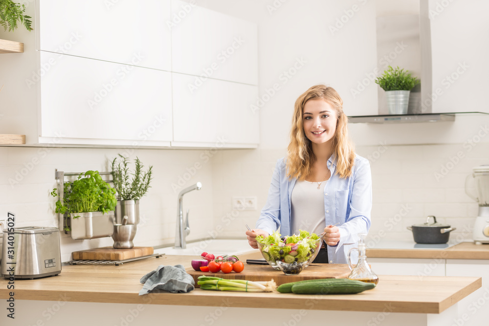 Young happy blonde girl preparing healthy salad in home kitchen