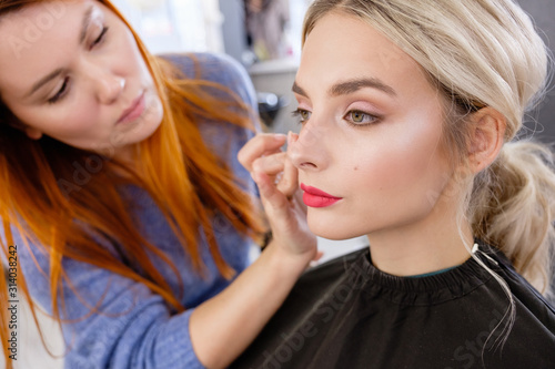 Young red-haired woman make-up artist puts make-up on face of beautiful blonde girl in beauty salon. Concept of personal care and celebration preparation. Daily self-care, beauty treatments in spa