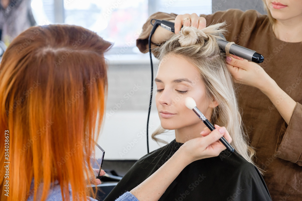 Fototapeta premium Women redhear make-up artist and hairdresser do hairstyle and stylish makeup for beautiful young blonde girl before start of fashion show. Colorful makeup design beauty and art salon concept