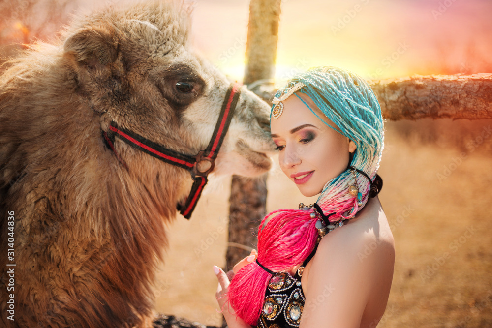 Obraz premium Beautiful girl with multi-colored braids in oriental clothes. Princess with a camel at sunset. in desert