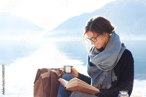 Young woman reading book at sea beach. Cozy winter picnic by morning mountains. Happy student in blue scarf enjoying traveling. Girl traveler drinks coffee. Solo female tourism. Lifestyle moment. photo