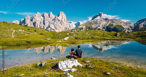 Awesome nature Landscape. Unknown couple sitting on green grass over amazing lake with turquoise water of in Tre Cime di Lavaredo national park. Wonderful Dolomites alps scenery.