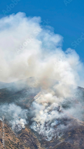 Vertical Aerial view of mountain with white smoke from wild forest fire on a sunny day
