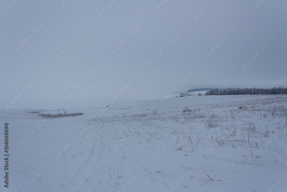 A snow-covered field and a large river covered with ice and snow merge into one white space at winter dusk in Russia