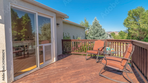 Pano Wooden balcony with metal armchairs facing the sliding glass door of a home