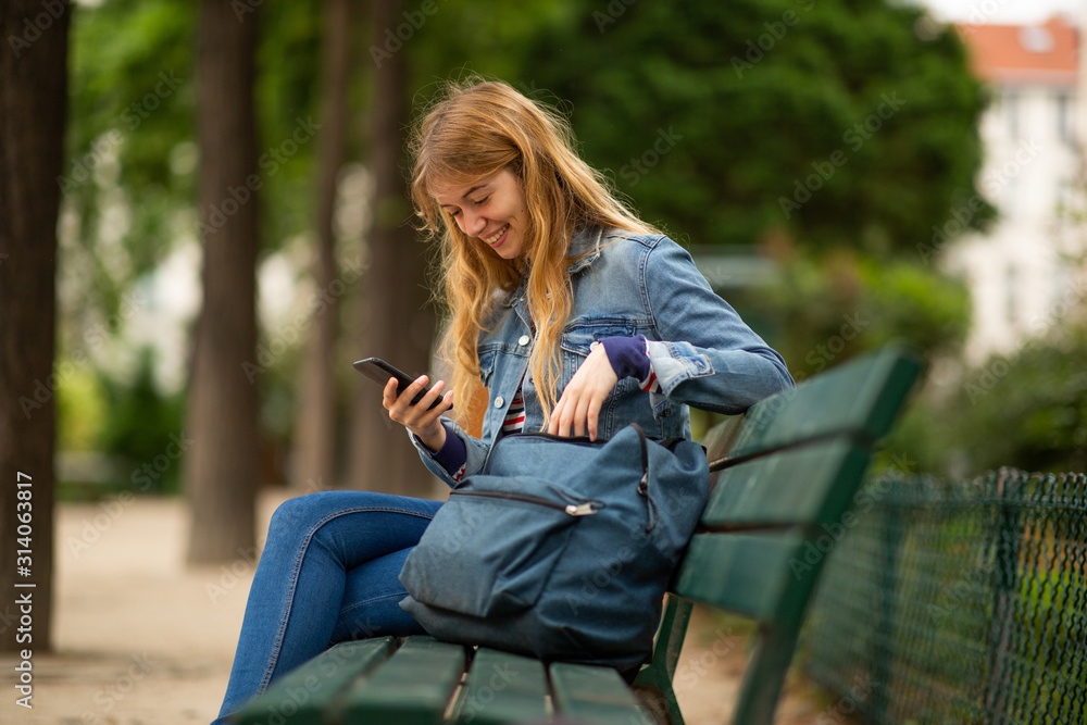 Side of smiling young woman sitting on park bench with mobile phone and bag Side of smiling young woman sitting on park bench with mobile phone and bag