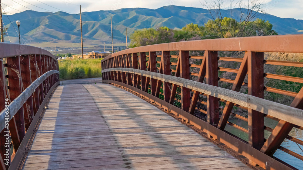 Fototapeta premium Pano Sunlit wooden deck and metail railing of bridge over lake with mountain view