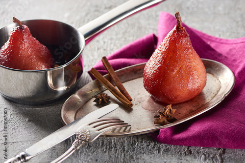delicious pear in wine with cinnamon and anise on silver plate and in stewpot on grey concrete surface with pink napkin, knife and fork