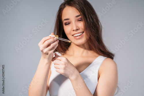 Beauty portrait of smiling young asian woman girl holding pipette with cosmetic oil near clean face on grey background, focus on hands