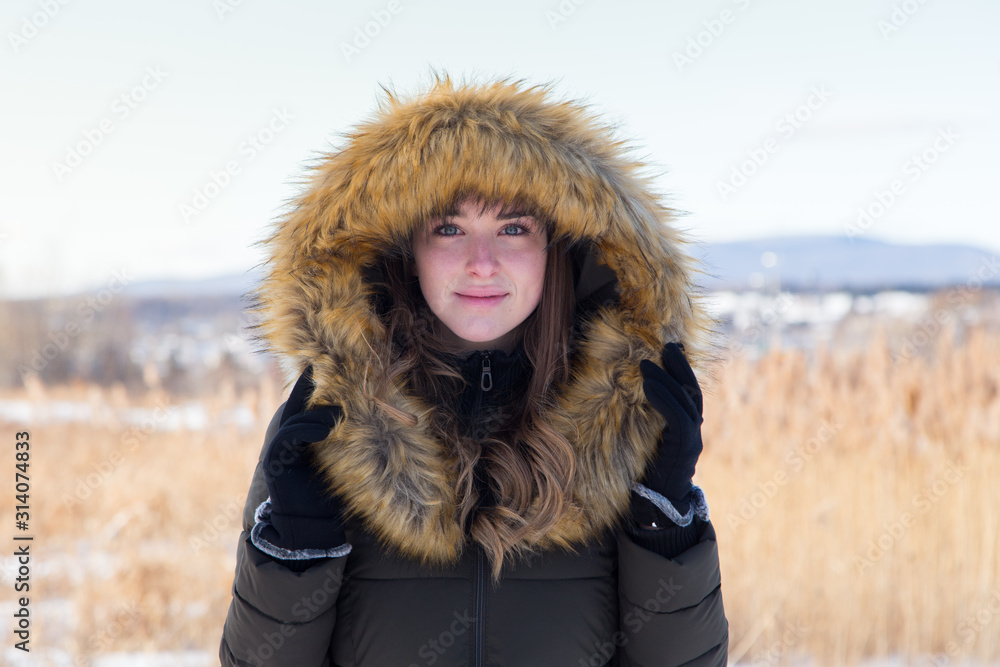 Medium horizontal portrait of pretty smiling blonde young woman in winter coat holding her faux fur trimmed hood up while standing outside in field during a sunny winter afternoon Medium horizontal portrait of pretty smiling blonde young woman in winter coat holding her faux fur trimmed hood up while standing outside in field during a sunny winter afternoon