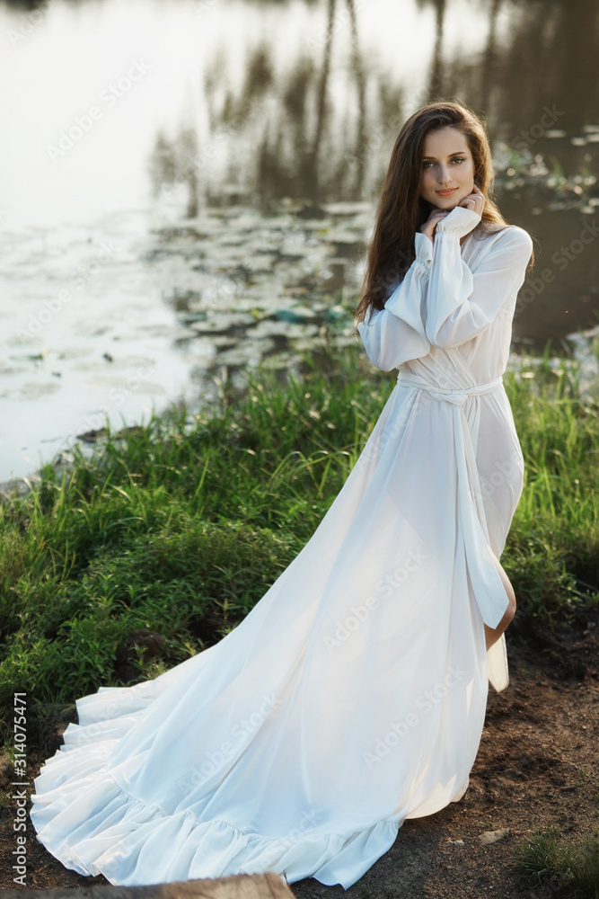 Beautiful slender woman in a long white dress walks in the morning near the lake. Brown-haired girl with long hair walks on the grass in the village, natural cosmetics and makeup