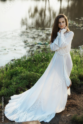 Beautiful slender woman in a long white dress walks in the morning near the lake. Brown-haired girl with long hair walks on the grass in the village  natural cosmetics and makeup
