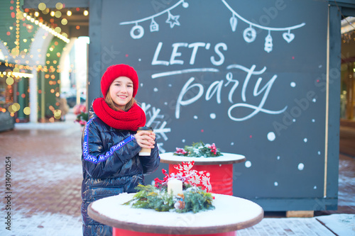 Smiling teenage girl in red hat enjoy cup of hot beverage on Christmas fair. 