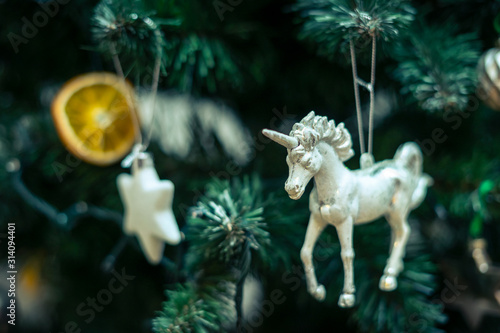 White unicorn as a Christmas tree ornament hanging on a Christmas tree in the company of baubles, an orange patch and white stars.