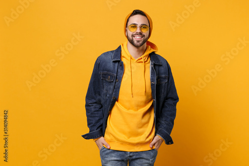 Smiling young hipster guy in fashion jeans denim clothes posing isolated on yellow orange background studio portrait. People emotions lifestyle concept. Mock up copy space. Holding hands in pockets.