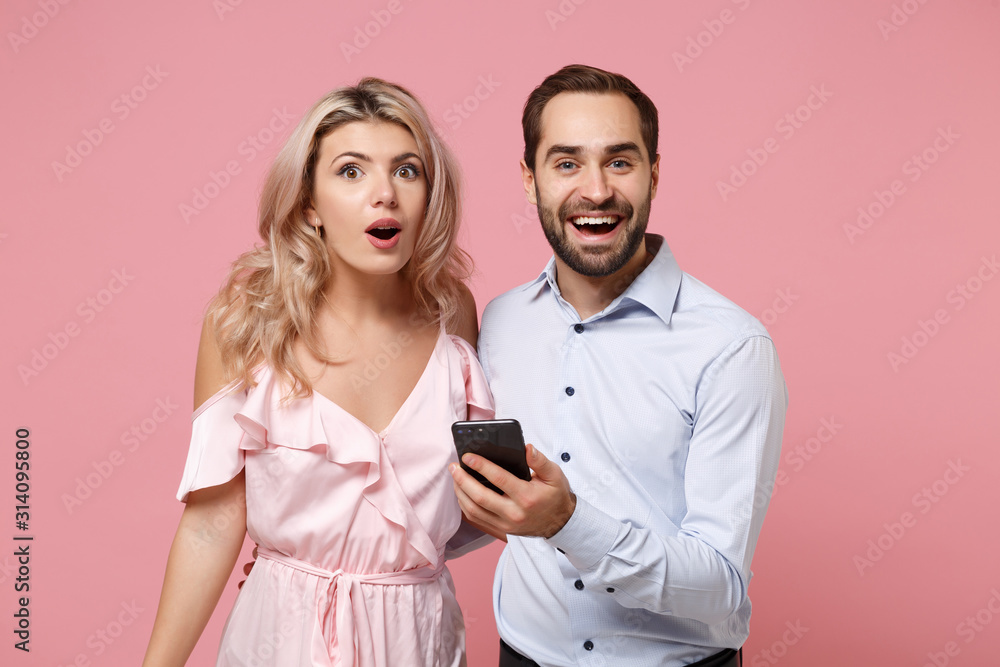 Shocked young couple two guy girl in party outfit celebrating isolated on pastel pink background. Valentine's Day Women's Day birthday holiday party concept. Using mobile phone, typing sms message. Shocked young couple two guy girl in party outfit celebrating isolated on pastel pink background. Valentine's Day Women's Day birthday holiday party concept. Using mobile phone, typing sms message.
