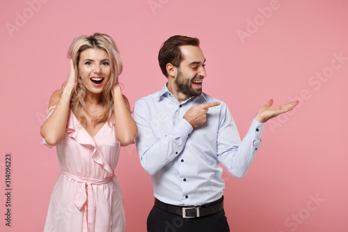 Cheerful young couple two guy girl in party outfit celebrating posing isolated on pastel pink background in studio. Valentine's Day Women's Day birthday holiday concept. Point index finger hand aside.